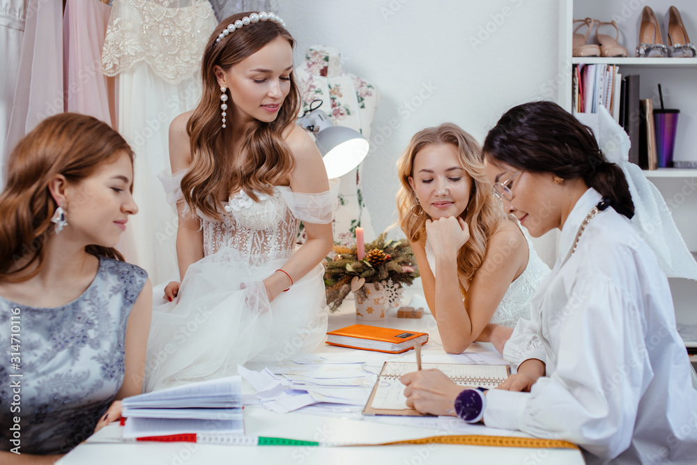 brunette adult woman designer communicate and explain which dresses are trendy, explain varieties, colors and style of dresses to young caucasian girls customers, models. isolated in wedding salon brunette adult woman designer communicate and explain which dresses are trendy, explain varieties, colors and style of dresses to young caucasian girls customers, models. isolated in wedding salon