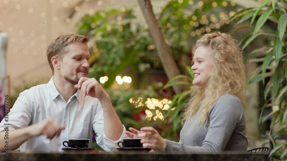Cheerful couple in casual clothes having joyful conversation while sitting at table and drinking coffee against blurred interior including much of green plants and bright lamps in contemporary cafe Cheerful couple in casual clothes having joyful conversation while sitting at table and drinking coffee against blurred interior including much of green plants and bright lamps in contemporary cafe
