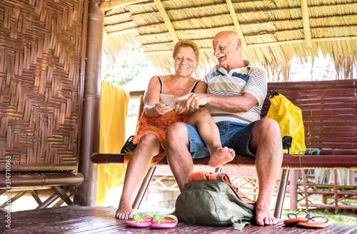 Happy senior couple using mobile smart phone at bungalow luxury resort - Active elderly and travel concept always connected with new trends and technology at adventure trip around world - Warm filter