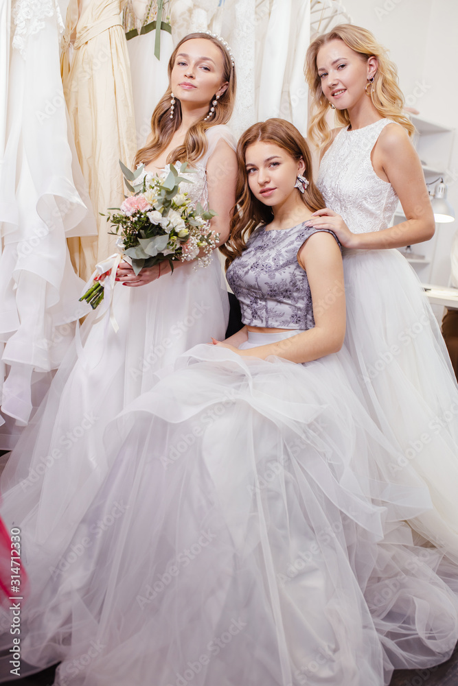 three beautiful young romantic ladies in trendy wedding dresses, future brides, going to be married, posing together at camera among dresses, holding flowers in hands three beautiful young romantic ladies in trendy wedding dresses, future brides, going to be married, posing together at camera among dresses, holding flowers in hands