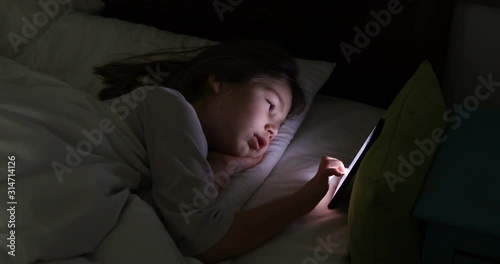 face lit of six years old girl typing on smart phone lying on bed at night  