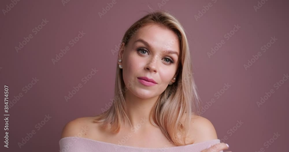 Young woman gently straightens hair behind an ear on pastel pink background. Blonde model posing and looking at camera. Gentle makeup on the face of model. Studio video session. Lifestyle. Copy space Young woman gently straightens hair behind an ear on pastel pink background. Blonde model posing and looking at camera. Gentle makeup on the face of model. Studio video session. Lifestyle. Copy space