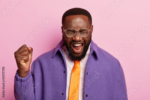 Pleased dark skinned man raises clenched fist, shouts yeah, celebrates success, wears orange tie, purple jacket, stands against pastel background, celebrates triumph, gets promotion in company