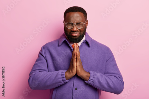 Hopeful black man keeps both hands pressed together in praying gesture, believes in good luck, wears optical glasses and purple jacket, isolated over rosy wall. Namaste gesture and meditation