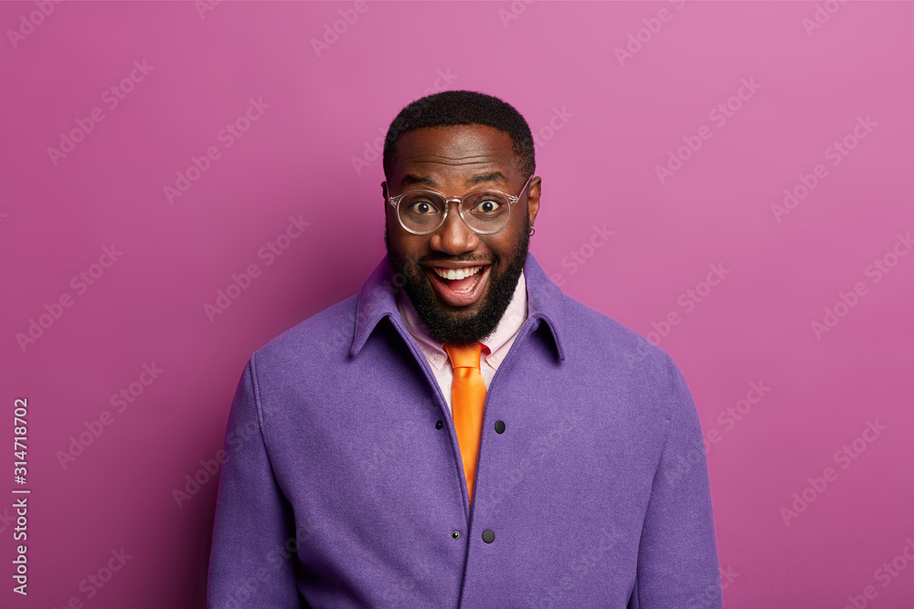 Friendly looking glad man laughs at camera, has funny conversation with interlocutor, wears transparent glasses, bright purple jacket, has toothy smile, white teeth, poses over lilac background Friendly looking glad man laughs at camera, has funny conversation with interlocutor, wears transparent glasses, bright purple jacket, has toothy smile, white teeth, poses over lilac background