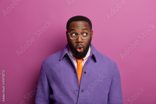 Surprised emotional bug eyed Afro American man with dark skin, keeps lips rounded, expresses shock after unexpected relevation, dressed in formal wear. People, facial expressions, lifestyle concept photo