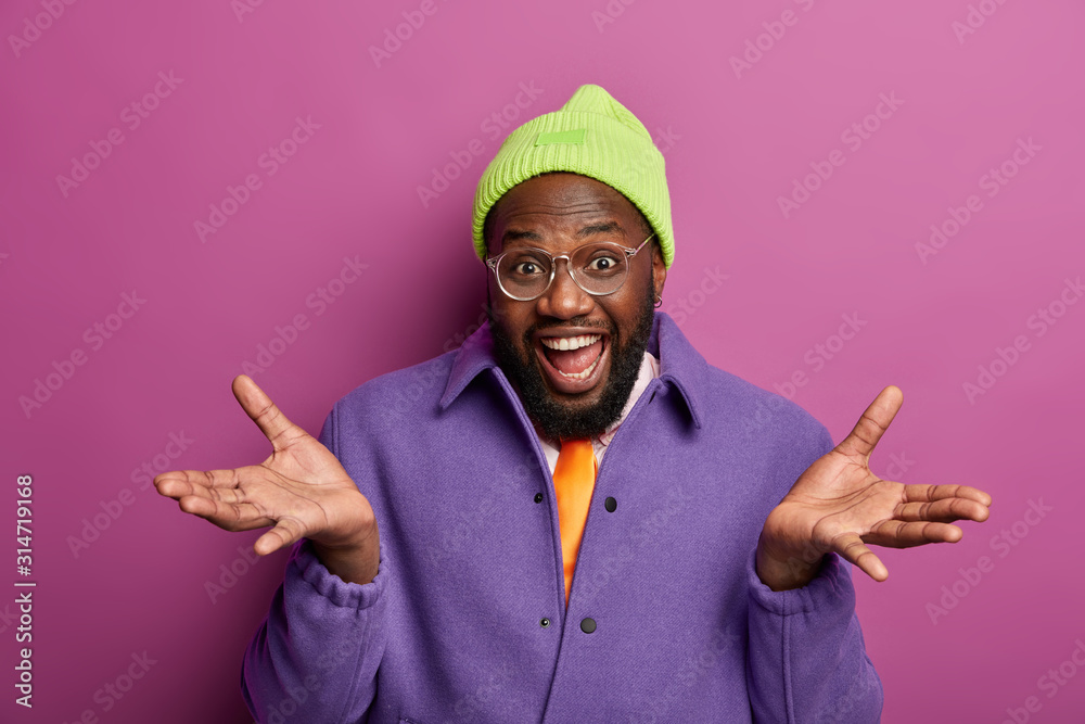 Photo of ethnic guy spreads palms, looks gladfully, chooses something, laughs sincerely, wears bright clothes, optical glasses, poses indoor, makes decision. So what are your suggestions to do?