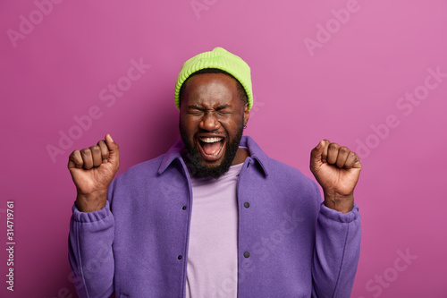 Emotional hipster cheers triumph, exclaims loudly, stands with clenches fists, dressed in fashionable clothing, expresses happiness, joy and excitement, stands indoor against bright vivid background photo