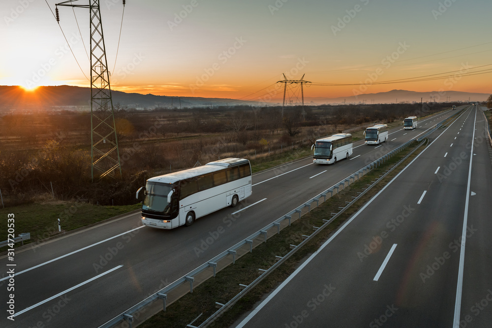 Caravan or convoy of Four white buses in line traveling on a country highway under amazing orange sunset sky. Highway transportation with white buses Caravan or convoy of Four white buses in line traveling on a country highway under amazing orange sunset sky. Highway transportation with white buses