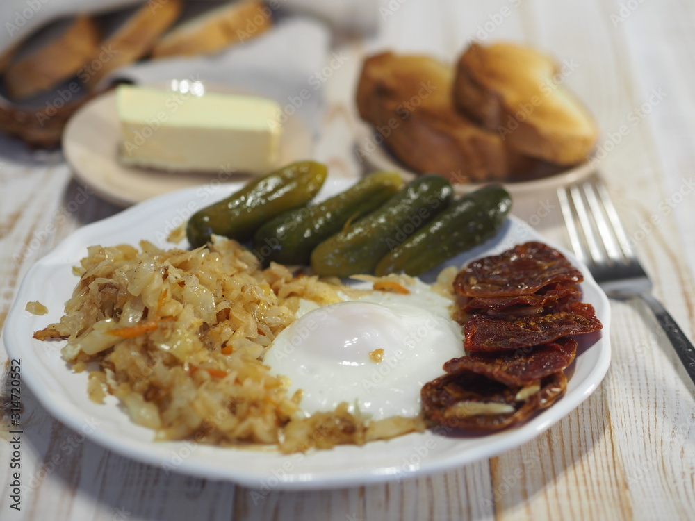 Home cooking. Fried cabbage, egg, pickles and dried tomatoes on a white plate on a wooden table. Bachelor breakfast.