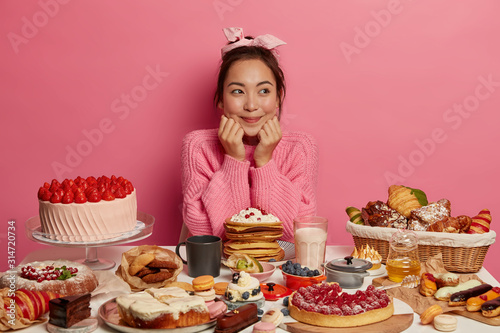 Photography Photo of satisfied Asian woman sits at table overloaded with many desserts, brea