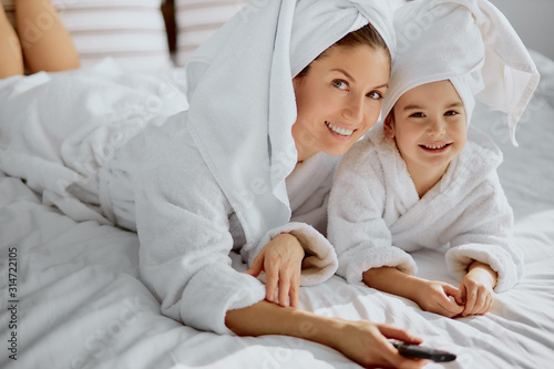 careful young caucasian woman and child in bathrobe and towel lie on bed holding tv remote and watch tv at free time
