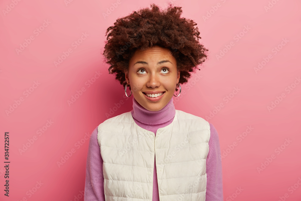 Positive woman with Afro curly haircut looks above with tender smile, wears turtleneck and white vest, notices something upwards, isolated over pink background. Happy mood, ethnicity, curiosity
