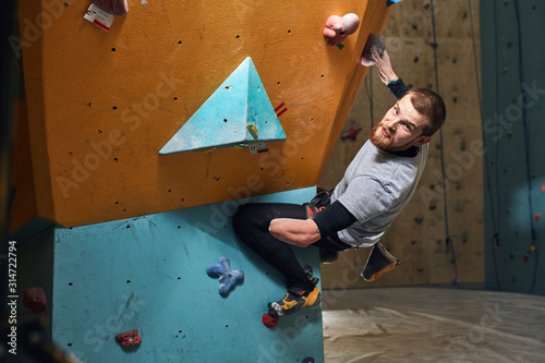 Athletic young male mountaineer trains hard despite his physical disability, enjoys his bouldering hobby, climbing difficult artificial wall after warming up exercises, overhanging on one hand.