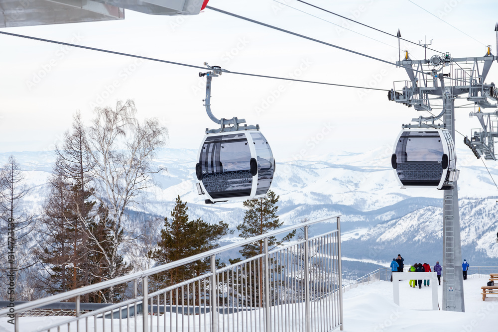 Obraz premium Landing place to a gondola cableway booth suspended on a cable where sits people with skis and snowboards on a background of mountains, trees with snow on winter sunset. Ski resorts and snowboarding.