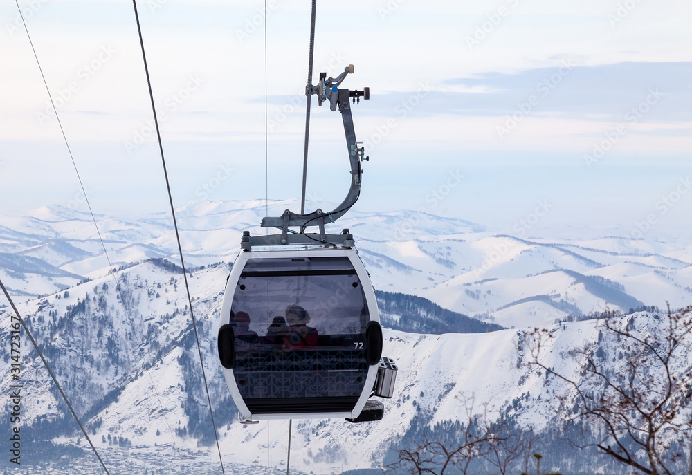 Obraz premium Cabin of a gondola cableway suspended on a rope where sits people with skis and snowboards high in the Altai mountains with snow and blue sky on winter sunset. Ski resorts and snowboarding.