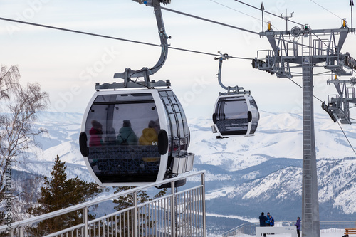 Landing place to a gondola cableway booth suspended on a cable where sits people with skis and snowboards on a background of mountains, trees with snow on winter sunset. Ski resorts and snowboarding.