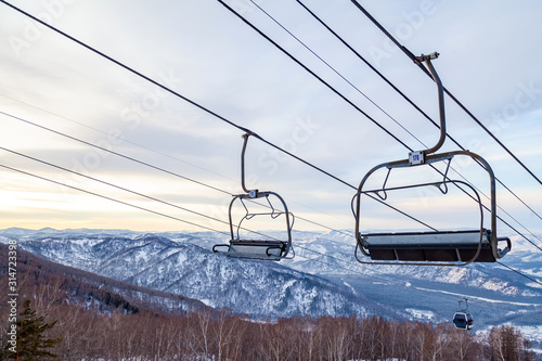 A ski lift chair cableway with a booth suspended on a cable in which sits people with skis and snowboards on a background of mountains, trees with snow on winter sunset. Ski resorts and snowboarding.