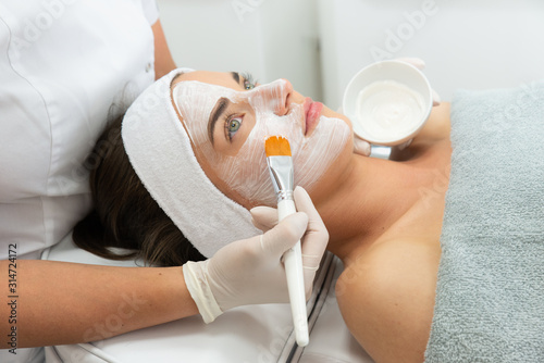 Kobieta w salonie kosmetycznym wykonuje zabieg oczyszczania cery. Kosmetyczka trzyma w dłoni pędzel i nakłada kwas na twarz kobiety.  photo