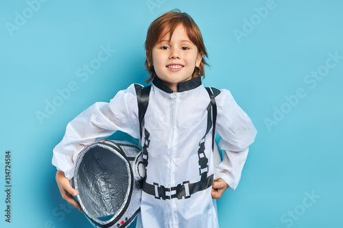 Portrait of cute caucasian boy wearing white protective suit and holding helmet stand isolated over blue background. look at camera photo