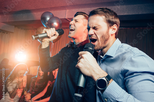 two young caucasian men in t-shirts singing in microphone in karaoke bar, having fun, celebrating. holiday, leisure, party concept photo