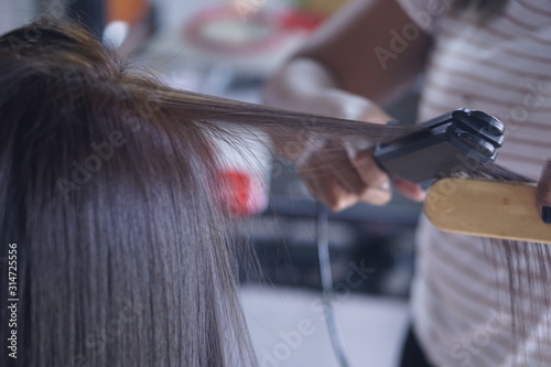 Hairstylist using curling iron of female client in hairdressing salon, closeup view. Hairdressing services. Creating hairstyle. Hair styling process.