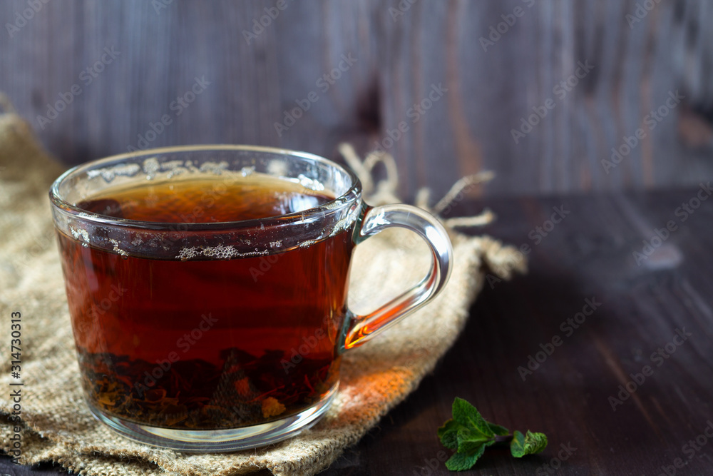 Cup of black tea with fresh green leaves of mint on a wooden table. Healthy lifestyle. Glass cup of black hot tea with sackcloth on a dark wooden background. Hot drinks. Cup of black tea with fresh green leaves of mint on a wooden table. Healthy lifestyle. Glass cup of black hot tea with sackcloth on a dark wooden background. Hot drinks.