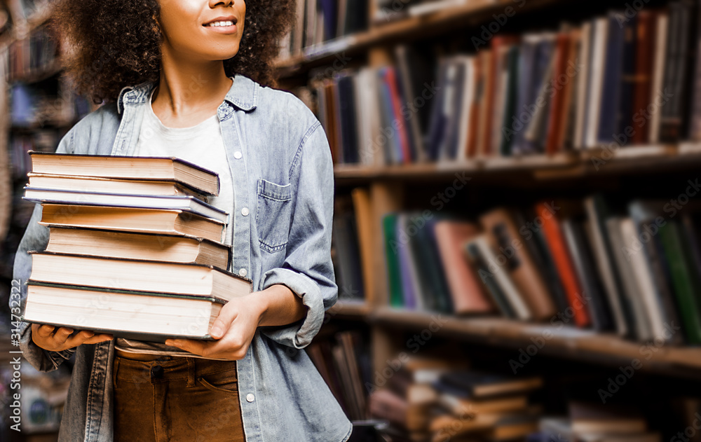 Photo for background, place for insertion, African American woman student holding a heavy armful of books in her hands against the background of bookshelves in a library. Learning concept, lifestyle - obrazy, fototapety, plakaty 