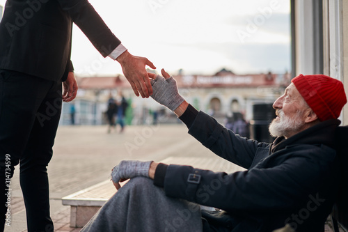 Senior beggar sitting beside street with request help with receive some coins from kind business man. Man hold out hand to homeless.