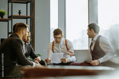young caucasian business team coworking analyzing and commenting reports sitting at table together, wearing formal clothes. isolated in modern boardroom, big panoramic window in the background