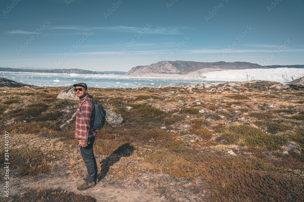 A young man traveler tourist standing in front of Eqip Sermia glacier called Eqi Glacier. Wall of ice in background. The concept of global warming and professional guides. On a sunny day with blue sky