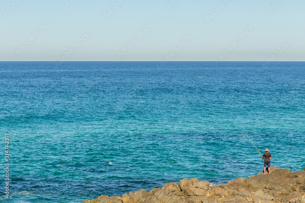 Beautiful blue ocean, a single fisherman stands on rocks alone with small blue waves