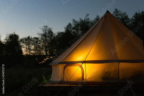 Illuminated yellow camping bell tent at night. Glamping bell tent glows at forest