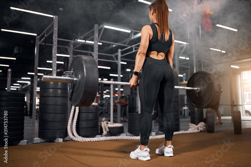 Fototapeta Naklejka Na Ścianę i Meble -  Tall sporty woman training in modern fitness studio, standing in front of professional barbell with back towards camera, spending evening in gym, getting strength to lift heavy barbell up, back view