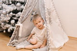 A small cute baby boy is sitting in the children's room in a tent Lodge wigwam next to the Christmas snow tree