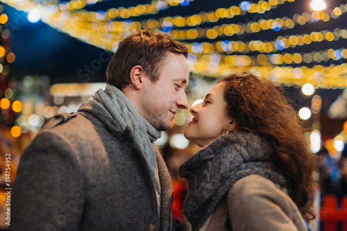 Romantic date for Valentine's day Outdoors. Couple in love having fun on christmas decoration lights street. Young man and Woman woman kiss and hug. Winter wonderland city  scene. New Year party