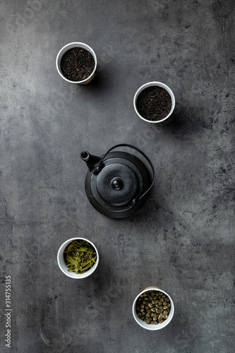 Black teapot and four different teas in paper cups on dark grey background