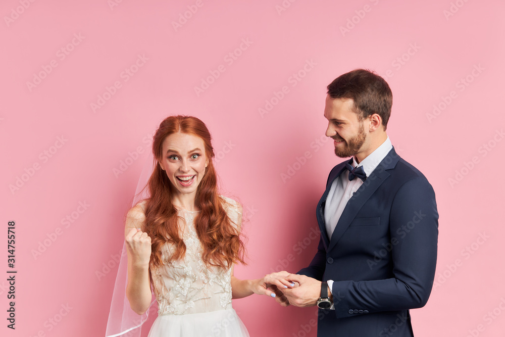 Marriage proposal. Redhaired woman get ring by handsome caucasian man in tuxedo putting ring on her finger. Isolated over pink background