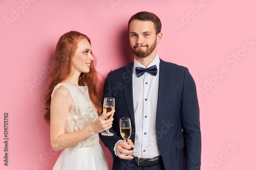 Beautiful woman and handsome na in tux with bow celebrate their wedding day together holding champagne, isolated over pink background photo