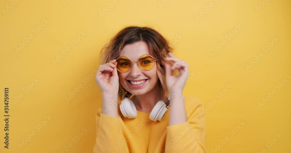 Portrait happy smiling young sexy girl in white headphones listens to music dancing, straightens yellow glasses on isolated yellow background. Pleasure. Emotions of people. Monotone. Copy space