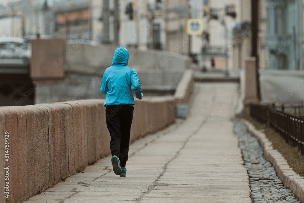 The girl is engaged in sports, jogging on the embankment. A woman in a blue jacket runs around the city.