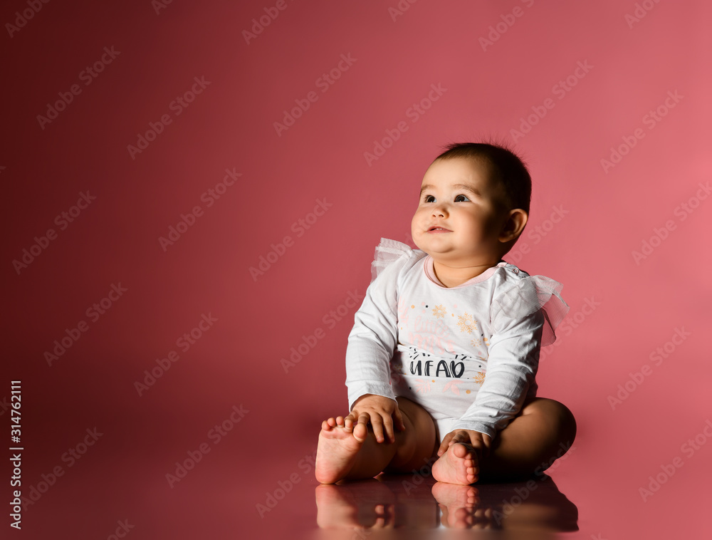 Little child in smart white bodysuit with inscription, barefoot. She smiling, sitting on floor against pink background. Close up Little child in smart white bodysuit with inscription, barefoot. She smiling, sitting on floor against pink background. Close up