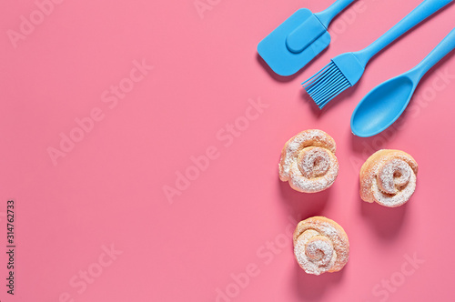 Homemade baked buns in form of rose spangled of powdered sugar near confectionery accessories rubber brush, spoon, spatula lies on pink background. Space for text. Top view