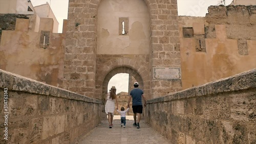 Mother and father are holding their baby boy hands and helping him to make first steps on an old cobbled street at a medieval castle in Spain in Bunyol or Bunol near Valencia. Young family on vacation