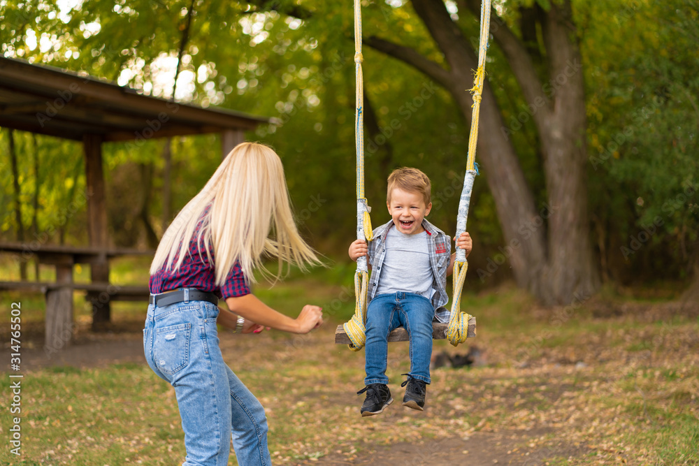 Young blonde mom shakes her little son on a swing in a green park. Happy childhood.