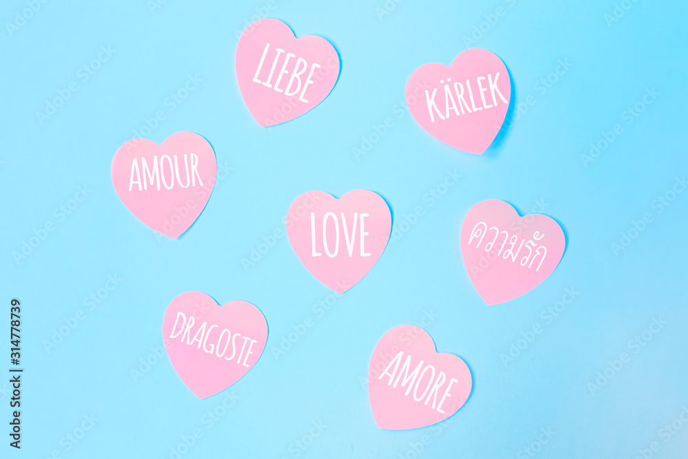 Some heart-shaped pink sticky notes with the word love written in different languages, such as English, Spanish, Italian, French, Dutch on a blue background. Valentines day, love concept.