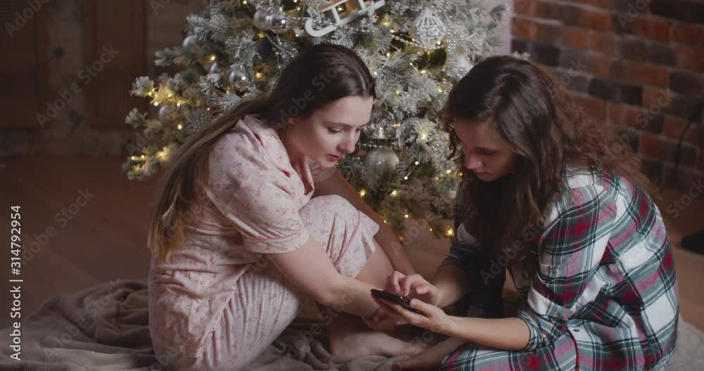 Two young women in pajamas are sitting near the New Year tree and viewing photos on the phone. Home cozy christmas concept Two young women in pajamas are sitting near the New Year tree and viewing photos on the phone. Home cozy christmas concept