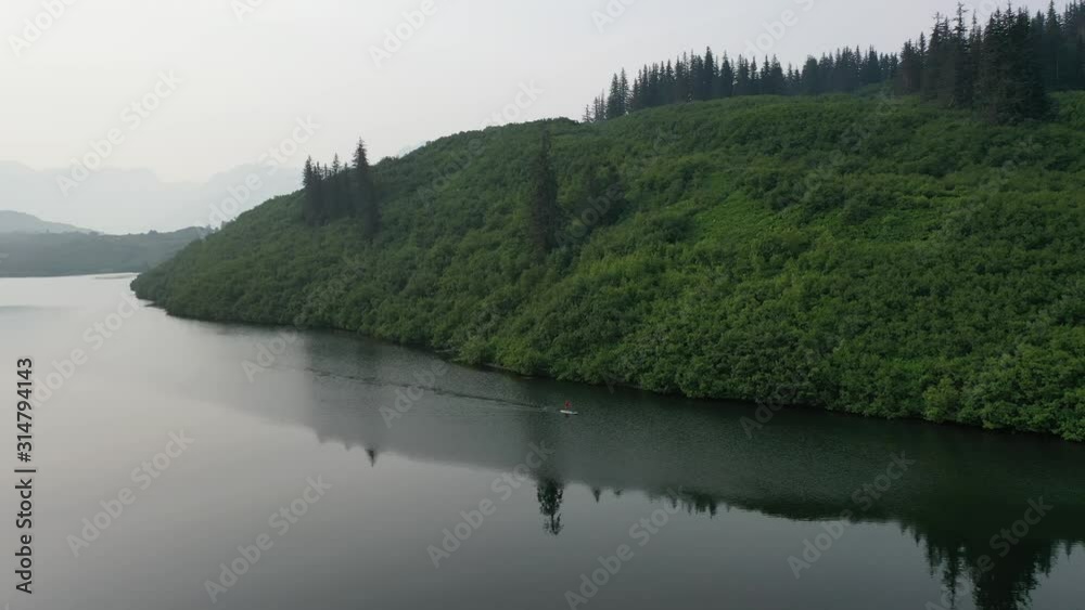 Slow zoom out and circling of paddle boarder on lake with vibrant green forest and mountain range. Slow zoom out and circling of paddle boarder on lake with vibrant green forest and mountain range.