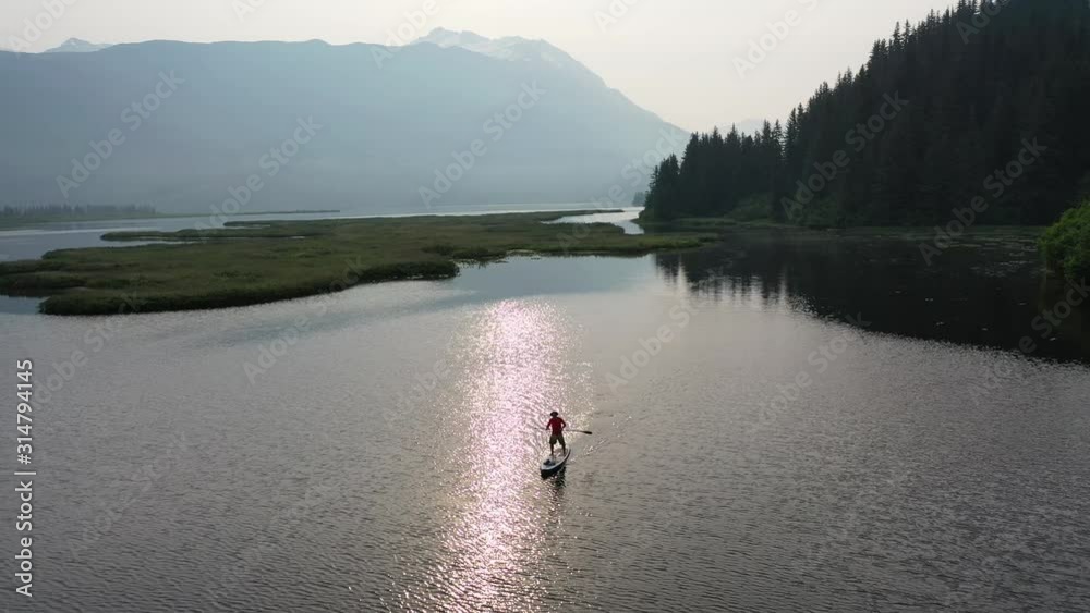 Paddle boarder holds up paddle while crossing lake with green forest and swamp behind him. Paddle boarder holds up paddle while crossing lake with green forest and swamp behind him.