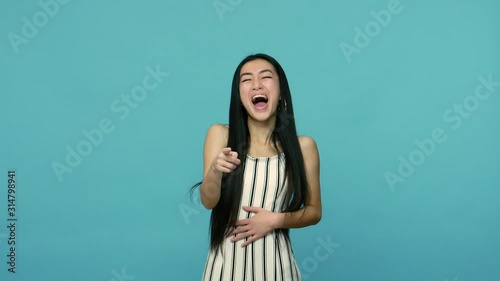 Extremely happy asian woman with long straight black hair in dress keeping hand on belly and laughing loudly and hysterically, pointing to camera, crazy laughter. indoor studio shot, blue background